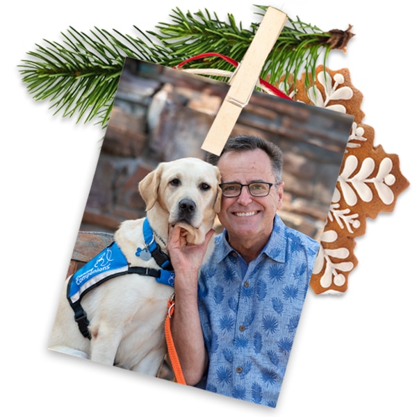 A smiling man with a yellow lab service dog wearing a blue vest - the image is pinned to evergreen with a gingerbread cookie behind