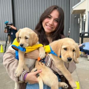 A smiling young woman holds two golden retriever puppies wearing yellow puppy vests